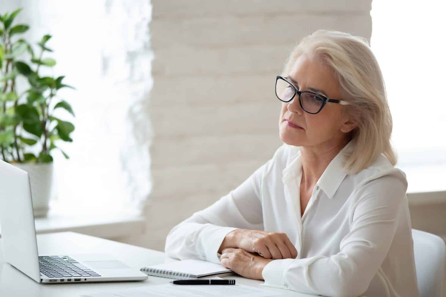 Thoughtful 60 years old businesswoman using laptop business vision. Stressful mature female employee working on financial report, sitting at office desk, manager solving business problem.