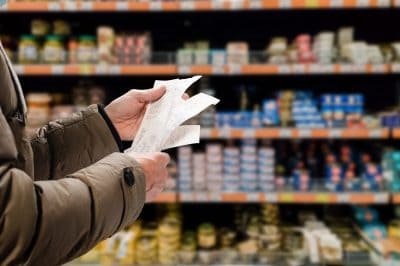 Close-up of a person's hands holding several white paper grocery receipts in a supermarket aisle. The person is wearing a brown jacket. In the background, out-of-focus shelves are stocked with various food products.