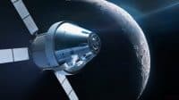 A highly detailed, silver-blue spacecraft, resembling an Orion capsule with its cylindrical service module and extended white solar arrays, is prominently featured in the foreground. It orbits the heavily cratered, dark grey surface of a celestial body, likely the Moon, which occupies the right half of the image. The spacecraft and the Moon are illuminated by a bright light source from the right, casting reflections and highlighting surface details, all set against the dark void of space with subtle hints of distant stars.