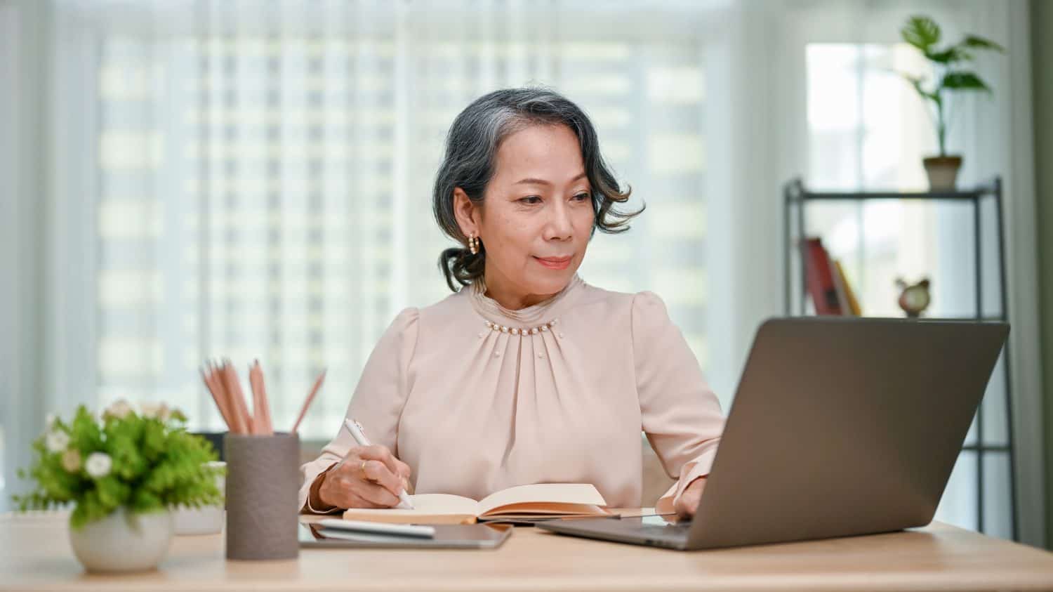 Charming 60s senior Asian female writer or businesswoman working at her desk, writing something on her book while using laptop computer.