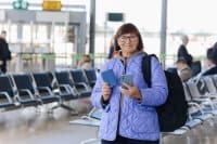 A smiling older woman with short brown hair and glasses stands in an airport terminal, holding a blue passport in her left hand and a grey smartphone in her right. She wears a quilted purple jacket over a grey hoodie and has a black backpack on her shoulder. The background is blurred, showing airport seating and other travelers.