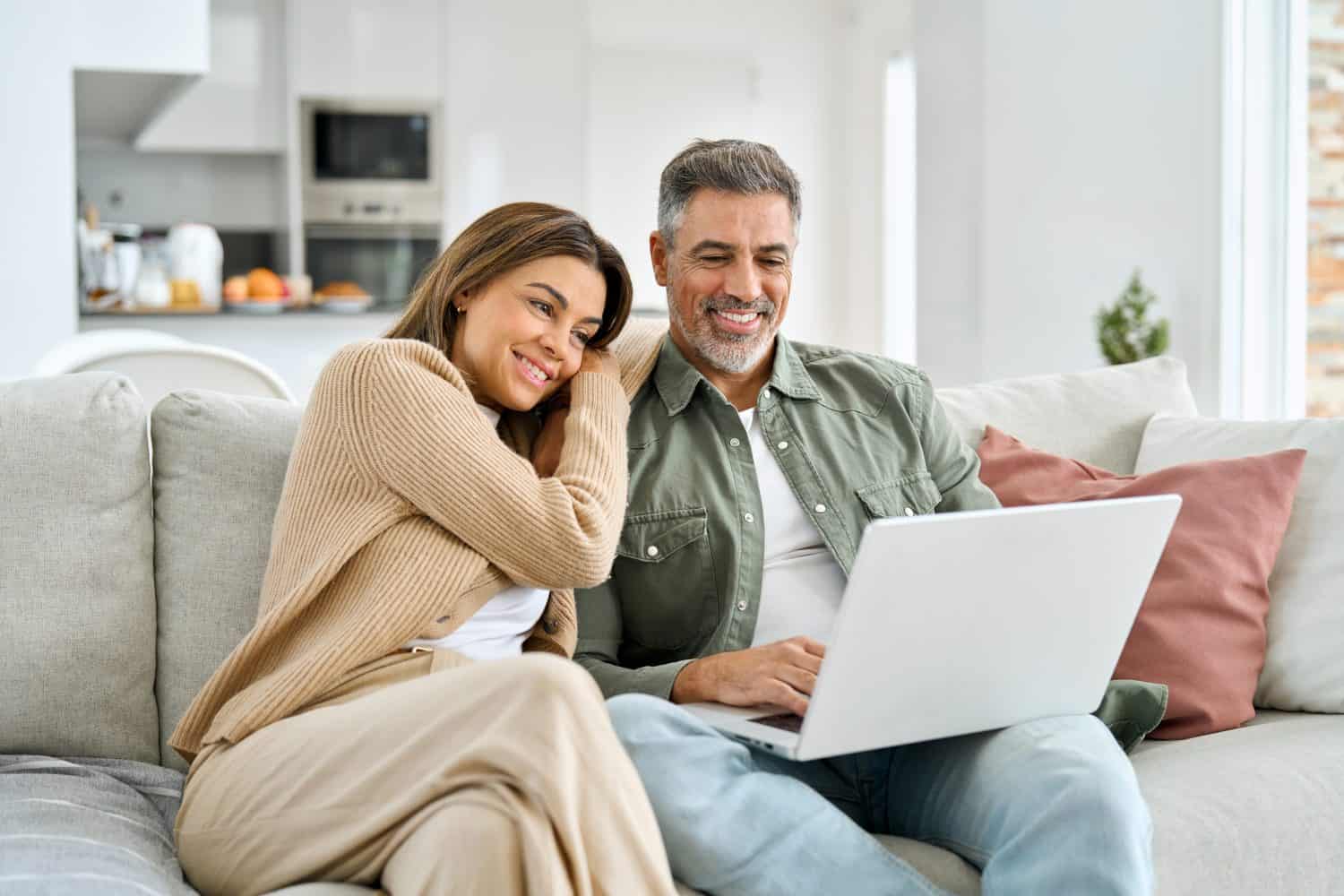 Happy middle aged couple using laptop relaxing on couch at home. Smiling mature man and woman looking at computer watching video, browsing or shopping in ecommerce store sitting on sofa in living room