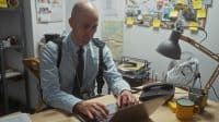 A bald man with a beard working on a laptop in a cluttered detective office surrounded by evidence boards.