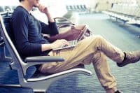 A man sits casually in an airport lounge chair, typing on a silver laptop resting on his lap. He wears a dark long-sleeved shirt and light brown pants, with his left hand on his chin, looking focused. A brown leather bag rests beside him, and rows of empty airport seating are visible in the soft-focus background.