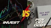 A split image visually contrasting 'INVEST' and 'PAY OFF DEBT'. The left side shows a dark screen with colorful stock market charts, including green and red candlesticks and various colored trend lines, with the word 'INVEST' in bold white letters at the bottom. The right side features a light gray surface with a spiral-bound notebook open to a page displaying 'PAY OFF DEBT' in purple text, accompanied by black-rimmed glasses and a pen. A large yellow 'VS' symbol on a red lightning bolt shape is centered in the middle, dividing the two concepts.