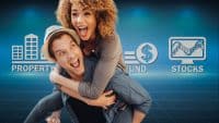 A man giving a piggyback ride to a woman, both smiling broadly with open mouths, are positioned in the foreground against a gradient blue background. In the background, from left to right, are three white graphic icons: a building with the text 'PROPERTY', a dollar sign with the text 'FUND', and a computer monitor displaying a stock chart with the text 'STOCKS'. The background features a subtle light blue grid pattern.