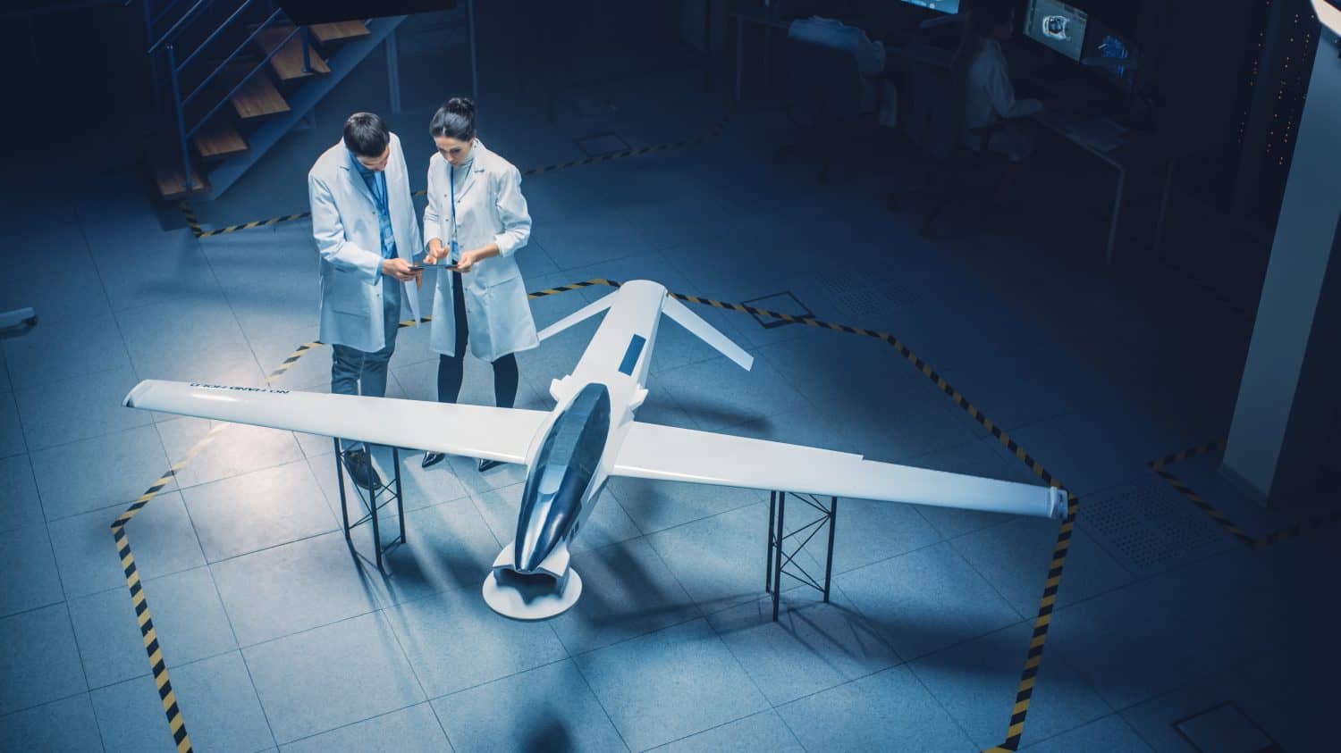 Two Aerospace Engineers Work On Unmanned Aerial Vehicle Drone Prototype. Aviation Scientists in White Coats Talking, Using Tablet Computer. Industrial Laboratory with Surveillance or Military Aircraft