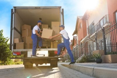 Two men in blue uniforms, caps, and white t-shirts are moving brown cardboard boxes into the back of a large white moving truck. One man is inside the truck, receiving a box from the other man who stands on the truck's ramp. The truck is parked on a paved road in front of houses with red tile roofs and black wrought-iron fences, under a bright blue sky with a prominent sun flare.