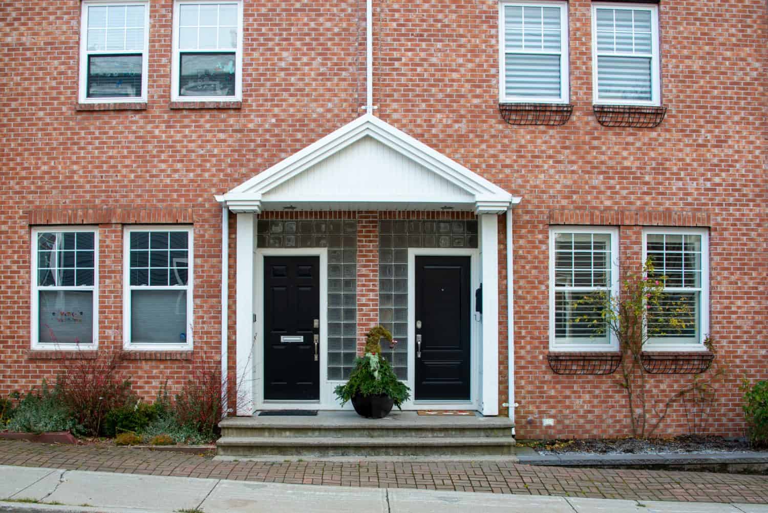 Two black metal doors with a Christmas gnome decoration on the steps of duplex houses. It has a red brick wall with white wooden trim. There are double hung windows on both sides of the entrance.