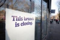 A close-up view of a white rectangular sign with purple text that reads 'This branch is closing,' affixed to the glass window of a building. The background, seen through and reflected in the window, is blurred, showing bare trees and a street scene with distant pedestrians under a bright sky.