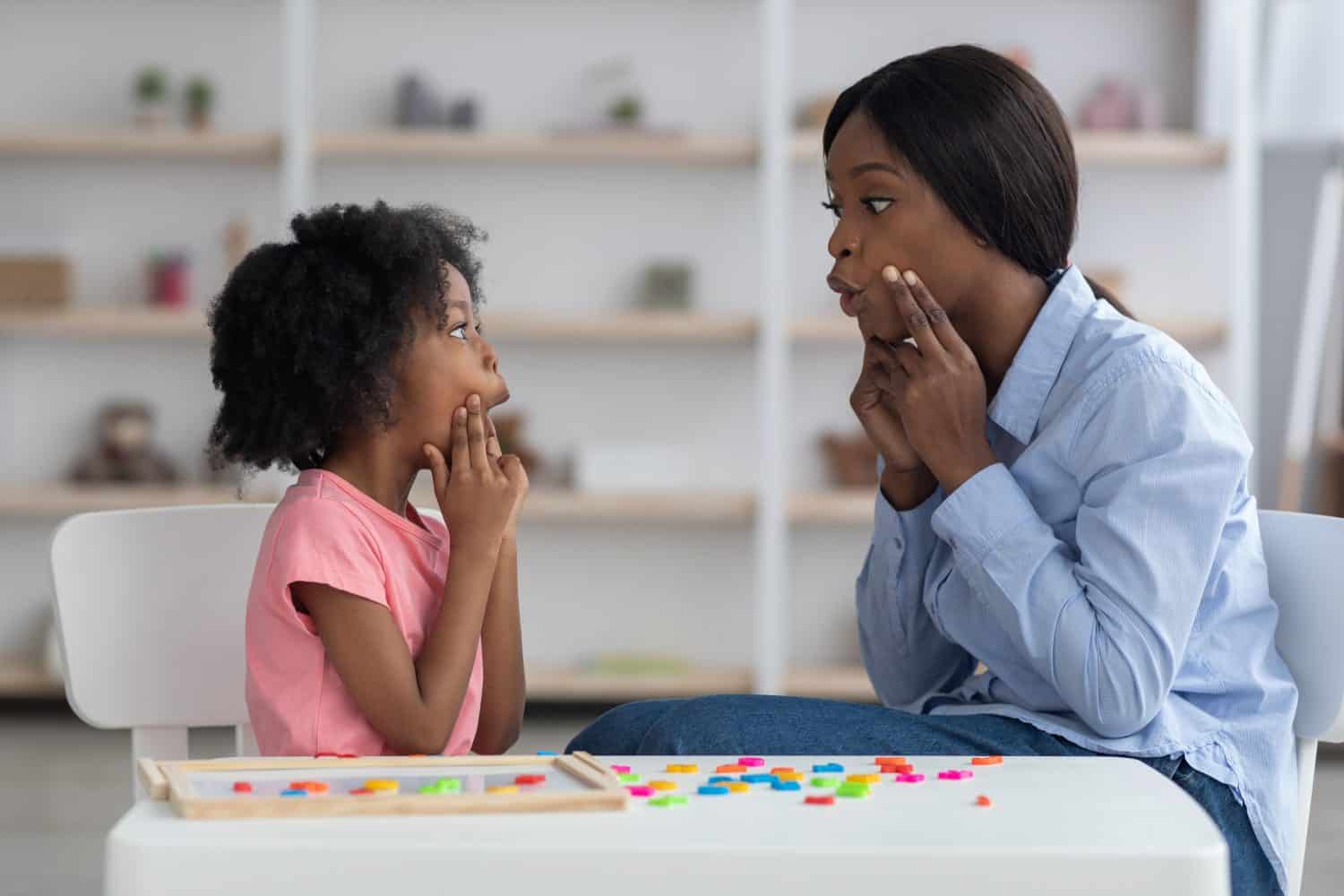 Young african american woman speech-language pathologist having lesson with little girl with cute curly hair, black teacher and pupil working on pronunciation, touching faces and grimacing