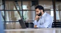 Thoughtful concentrated indian business man entrepreneur investor manager using computer, watching webinar working in office analyzing online data market thinking doing web research looking at laptop.