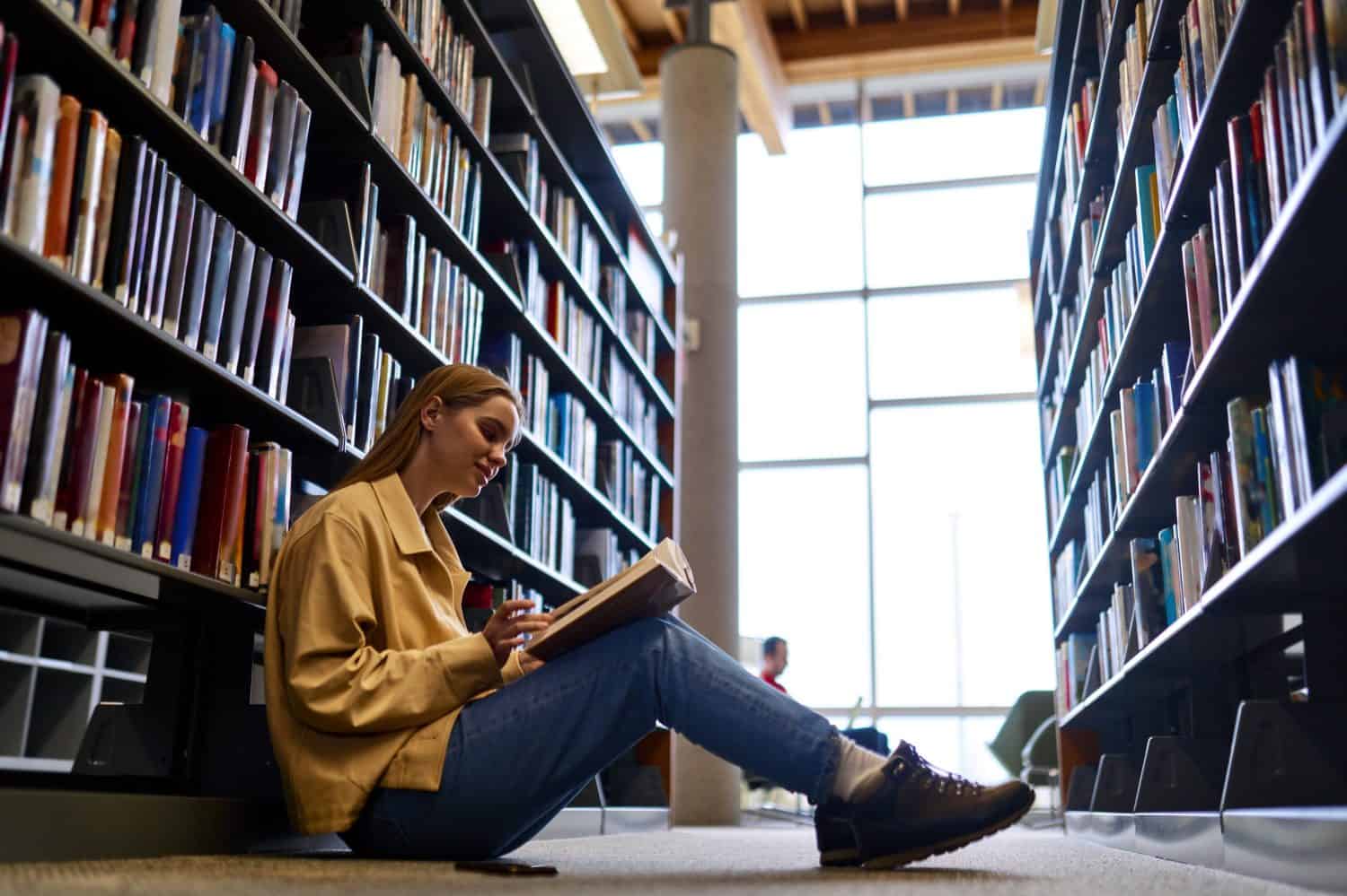 Smart pretty creative girl student holding book sitting on floor among bookshelves in modern university campus library or bookstore thinking of college course study reading literature doing research.