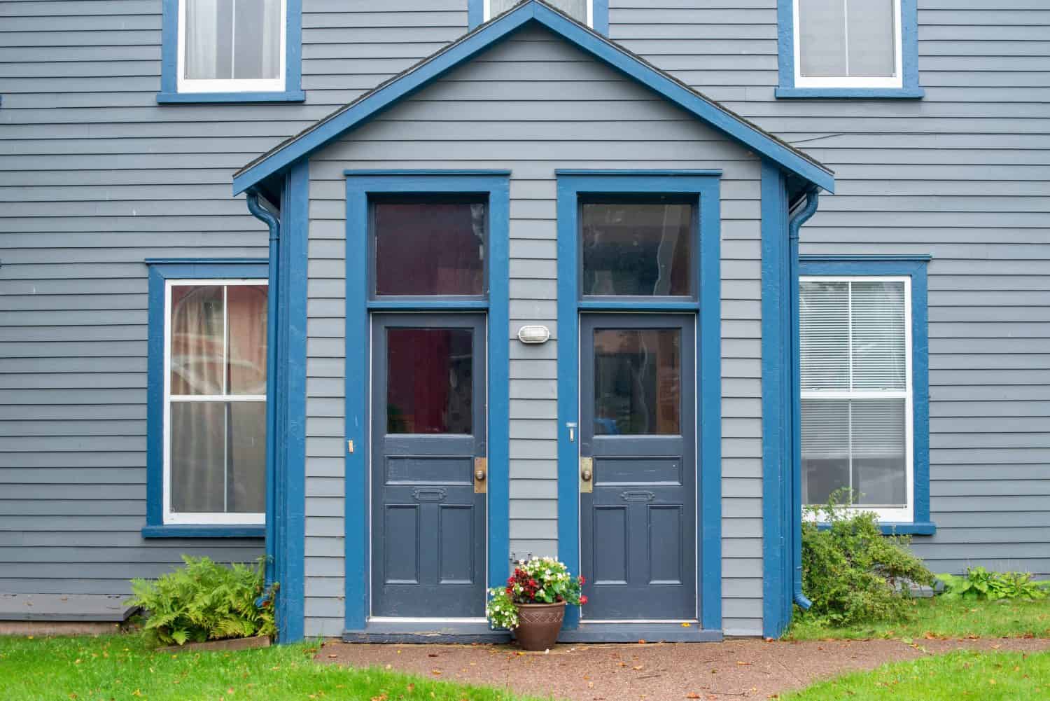 Two exterior half glass doors to a duplex. The building's wall is grey colored with royal blue trim and a pot of flowers in front. There are four double hung windows with curtains and blinds hanging.