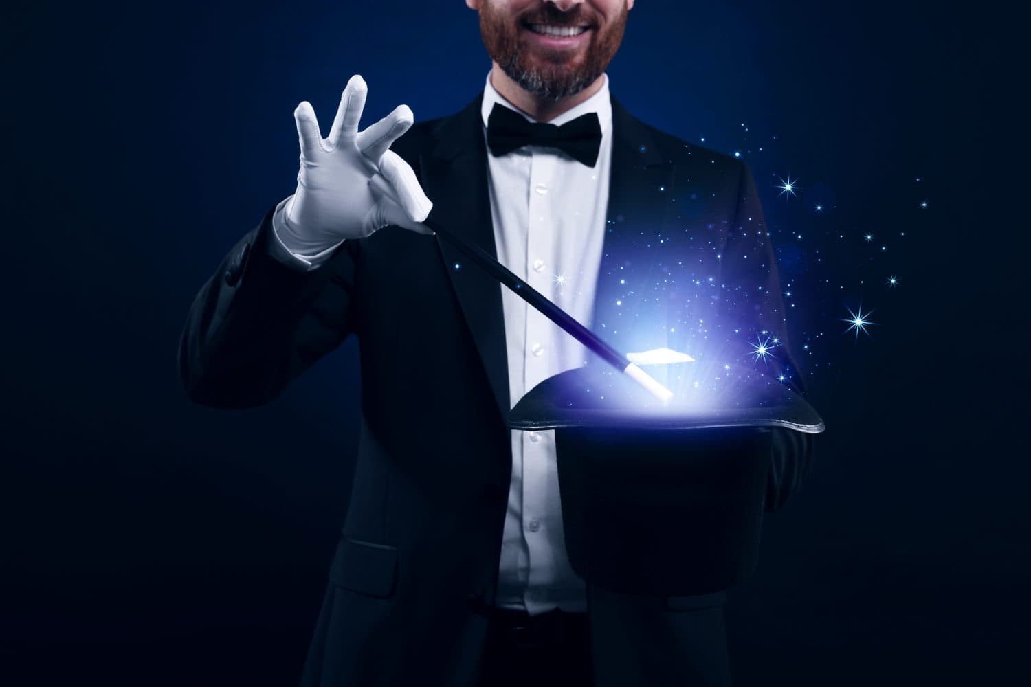 Smiling magician showing trick with wand and top hat on dark blue background, closeup