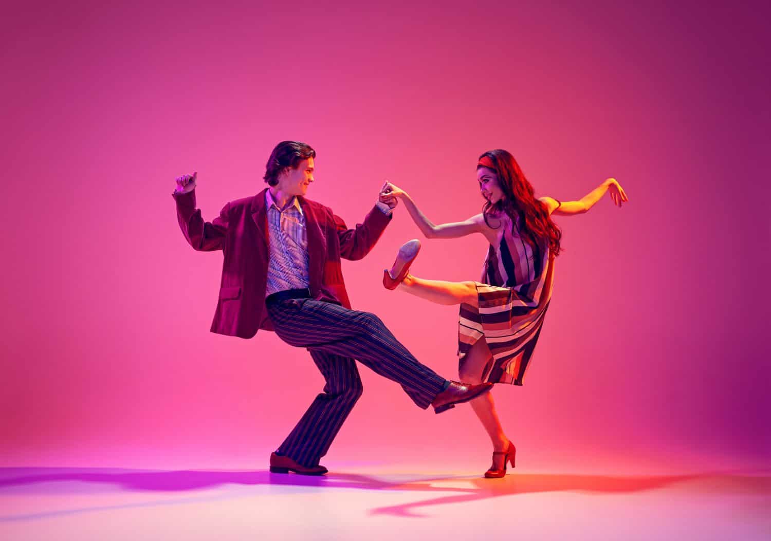 Stylish, beautiful young couple, man and woman in retro clothes dancing swing against pink background in neon light. Concept of hobby, dance class, party, 50s, 60s culture, youth