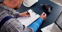 Overhead shot of a man in a plaid shirt and blue brace on his left forearm, writing on a 'Health Insurance Claim Form' with a black pen. A white hard hat, a black and blue power drill, and a partially visible white laptop are on the dark blue desk beside him.