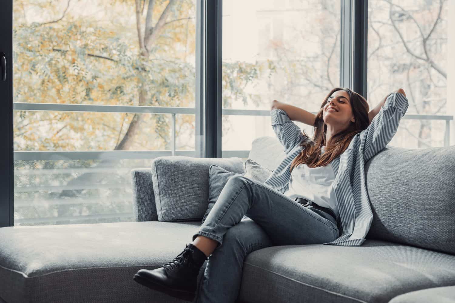 Relaxed serene pretty young woman feel fatigue lounge on comfortable sofa hands behind head rest at home, happy calm lady dream enjoy wellbeing breathing fresh air in cozy home modern living room