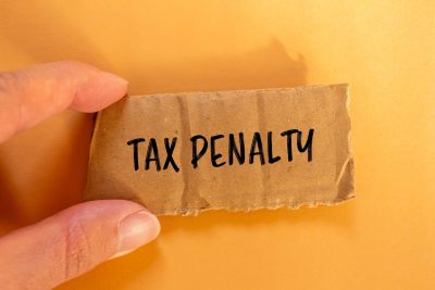 A hand holds a jagged piece of brown cardboard with the words 'TAX PENALTY' written in black marker. The background is a plain, warm yellow-orange color, with soft shadows cast by the hand and cardboard.