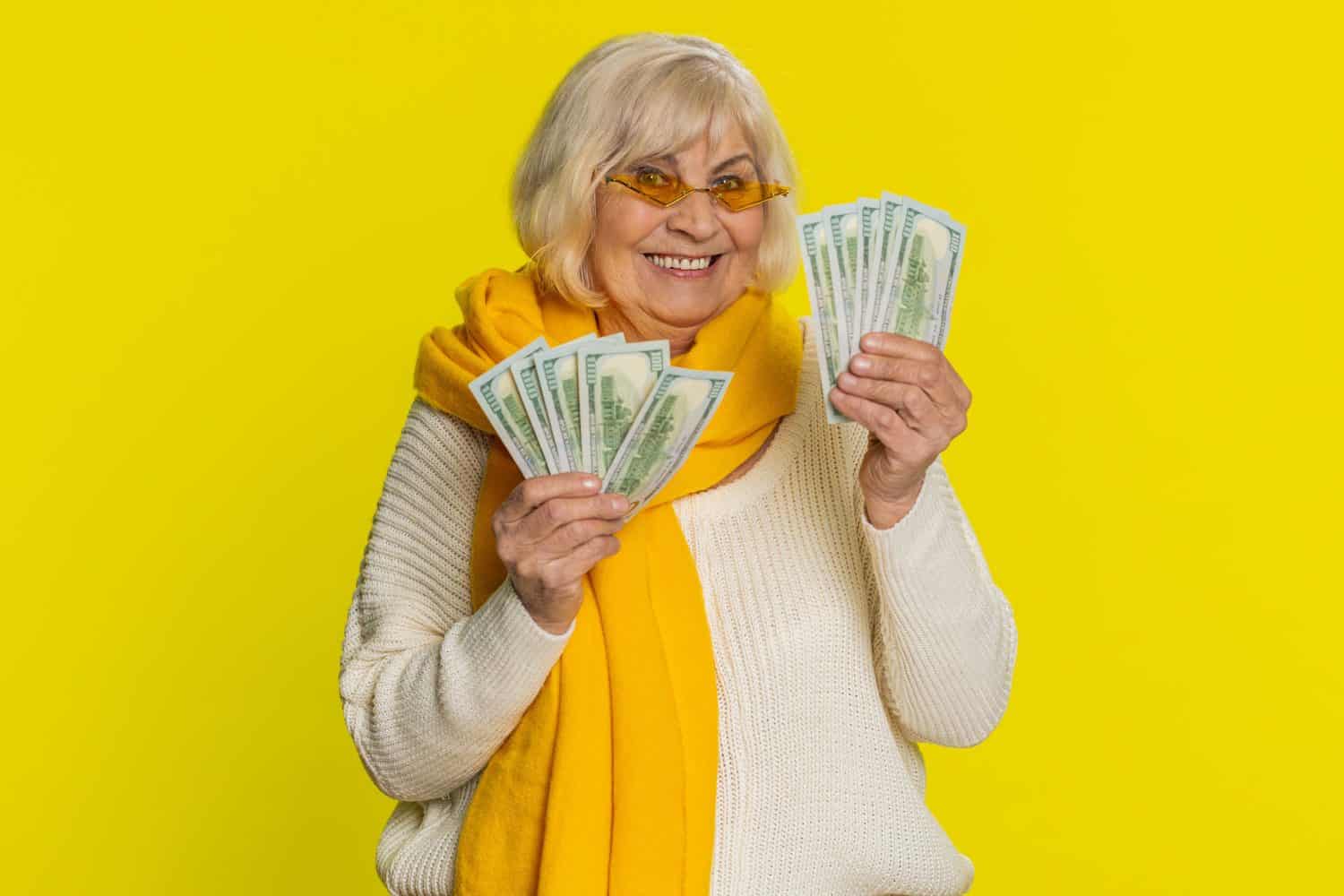 Senior old woman holding fan of cash money dollar banknotes celebrate dance success business career, lottery game winner, big income, wealth pension. Elderly grandmother pensioner on yellow background