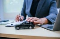 A close-up shot of a person in a dark suit sitting at a desk. Their left hand gently rests on a black toy SUV model car. Their right hand holds a pen, writing on a document or notebook. A silver laptop is visible on the right side of the desk, and the background shows a blurred office setting with blinds.