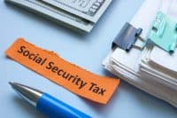 An orange slip of paper with 'Social Security Tax' written in black capital letters lies on a light blue surface. To the left is the tip of a blue and silver ballpoint pen. In the upper background, stacks of U.S. dollar bills are visible, and to the right, stacks of white paper are held together with a dark gray and a teal binder clip.