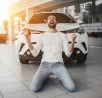 Yes, that's my new car! Customer in car dealership. Young man knelt down with keys in hand.