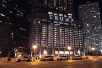 A wide night shot of the illuminated "TRUMP" sign on a tall, modern skyscraper with a curved facade. Numerous windows on the building are lit with warm white light. To the left, a darker, multi-windowed building rises. To the right, another white, multi-windowed skyscraper is visible. In the foreground, four white and blue "CHICAGO POLICE" SUVs are parked along a street, under bright yellow streetlights. A statue of a man pointing stands near the leftmost police car.