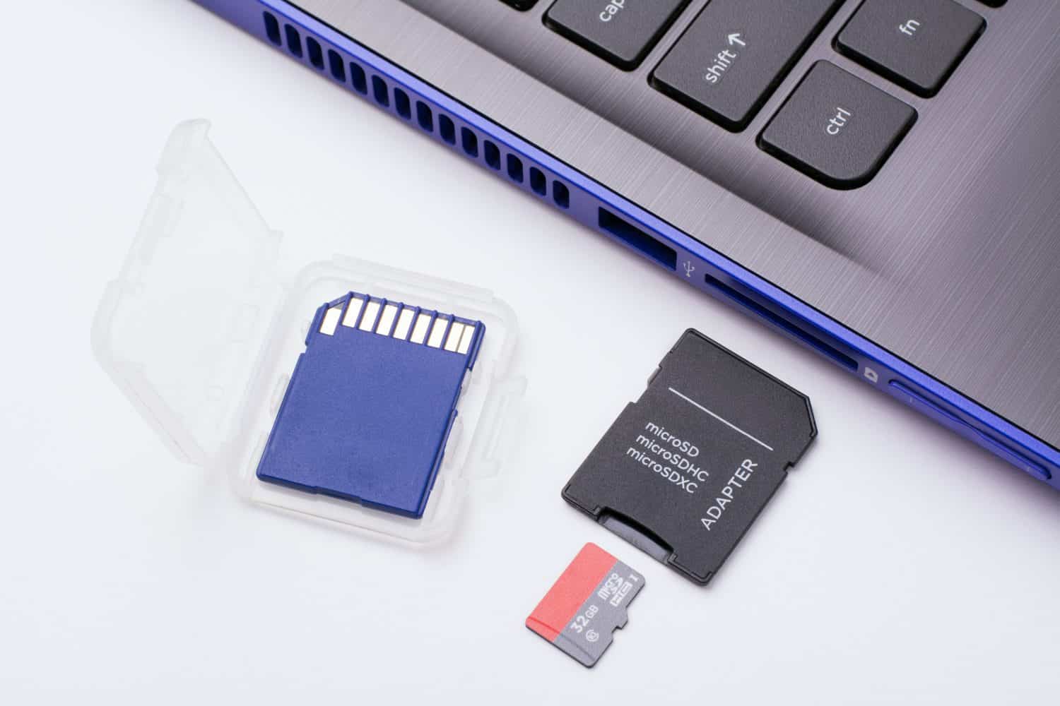 MicroSD memory card with adapter near the SD port of the modern blue laptop. One more blue SD memory card in a transparent case lays near the computer.