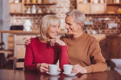 An older Caucasian couple sits at a wooden table in a cafe, looking at each other and smiling warmly. The woman, with blonde hair and a red sweater, holds one hand to her chin while the man, with grey hair and a brown sweater, has his arm around her shoulder. Two white coffee cups with saucers are on the table in front of them.