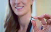 A close-up shot of a small, white, capsule-shaped device with a clear front tip, held between the thumb and forefinger of a person. The device is in sharp focus, while a smiling woman with long blonde hair is blurred in the background, suggesting a positive interaction with the object. The overall lighting is bright and clean.