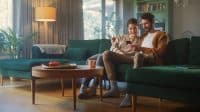 A young adult couple sits together on a dark green velvet sectional sofa in a sunlit living room. The woman, with short dark hair, smiles while holding a black smartphone, and the man, with a beard, has his arm around her shoulders and looks at the phone with a smile. A round wooden coffee table with a small tray and a light orange mug is in the foreground. A floor lamp with a beige shade stands next to the sofa, and a large houseplant is visible in the background near a window with light-colored curtains. The overall mood is relaxed and collaborative.