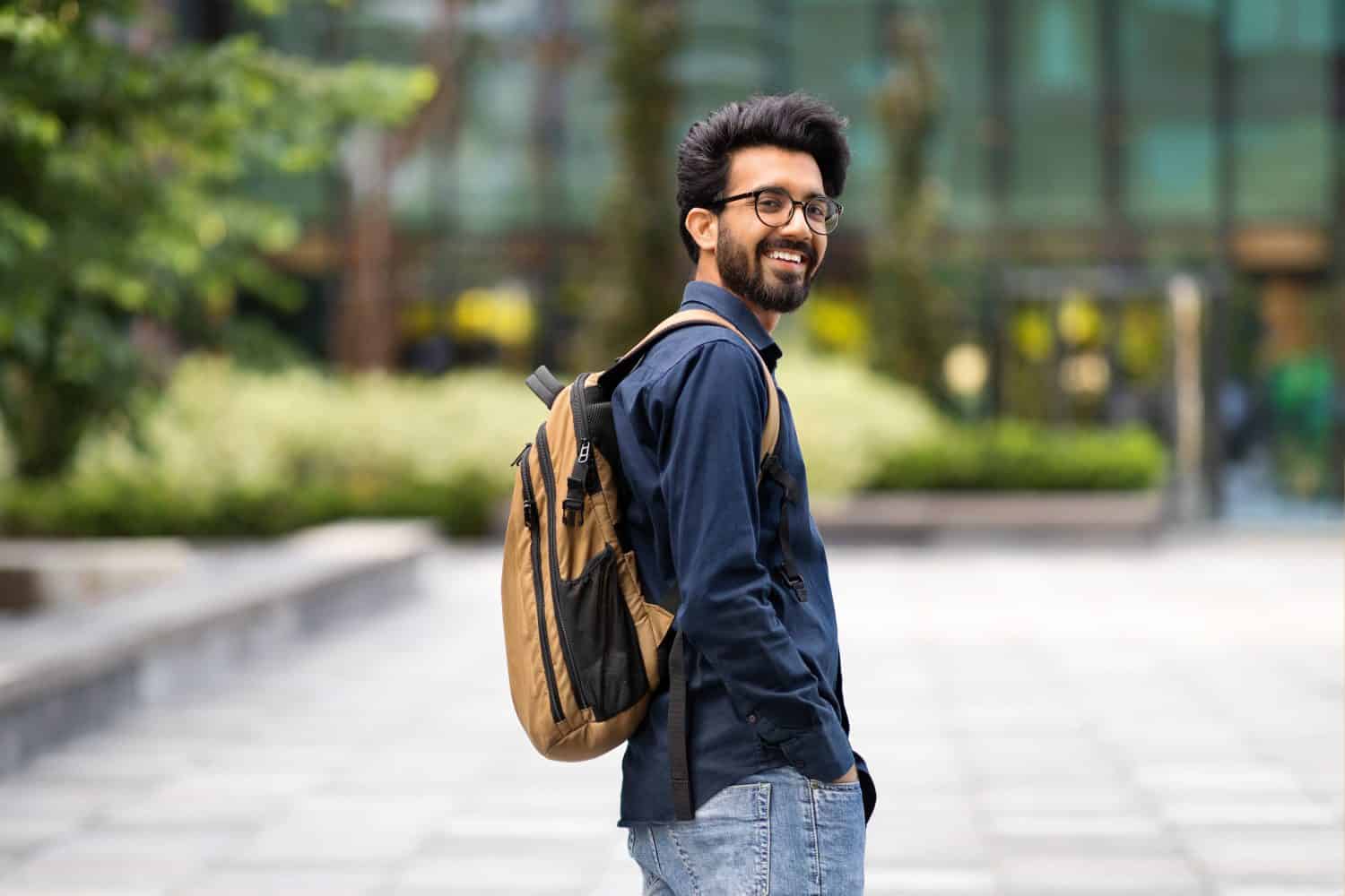 Cool cheerful young hindu guy wearing casual outfit with backpack walking by street, smiling at camera, going to job, copy space. Positive indian man student going to college, university