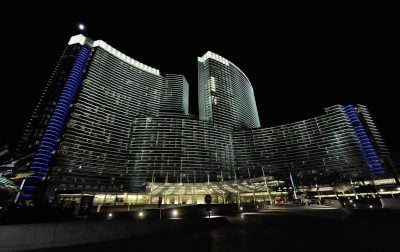 A wide-angle, low-angle night shot of a large, modern luxury hotel and casino complex with multiple towers, brightly lit. The buildings feature sweeping, curved facades with numerous windows showing warm interior lights and prominent vertical strips of vibrant blue accent lighting. A bright, full moon is visible in the dark sky above the highest point of the structure. The illuminated entrance and a roadway are visible at the base of the complex.