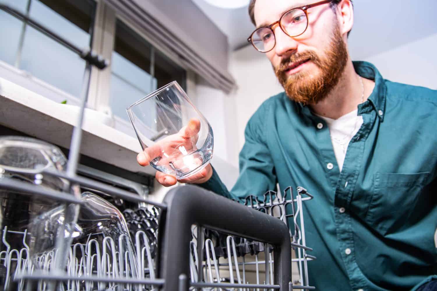Young adult man emptying ,loading, taking out a clean glass from a dishwasher. Household chores, duties with a modern kitchen appliance. An open dishwasher with clean glasses.