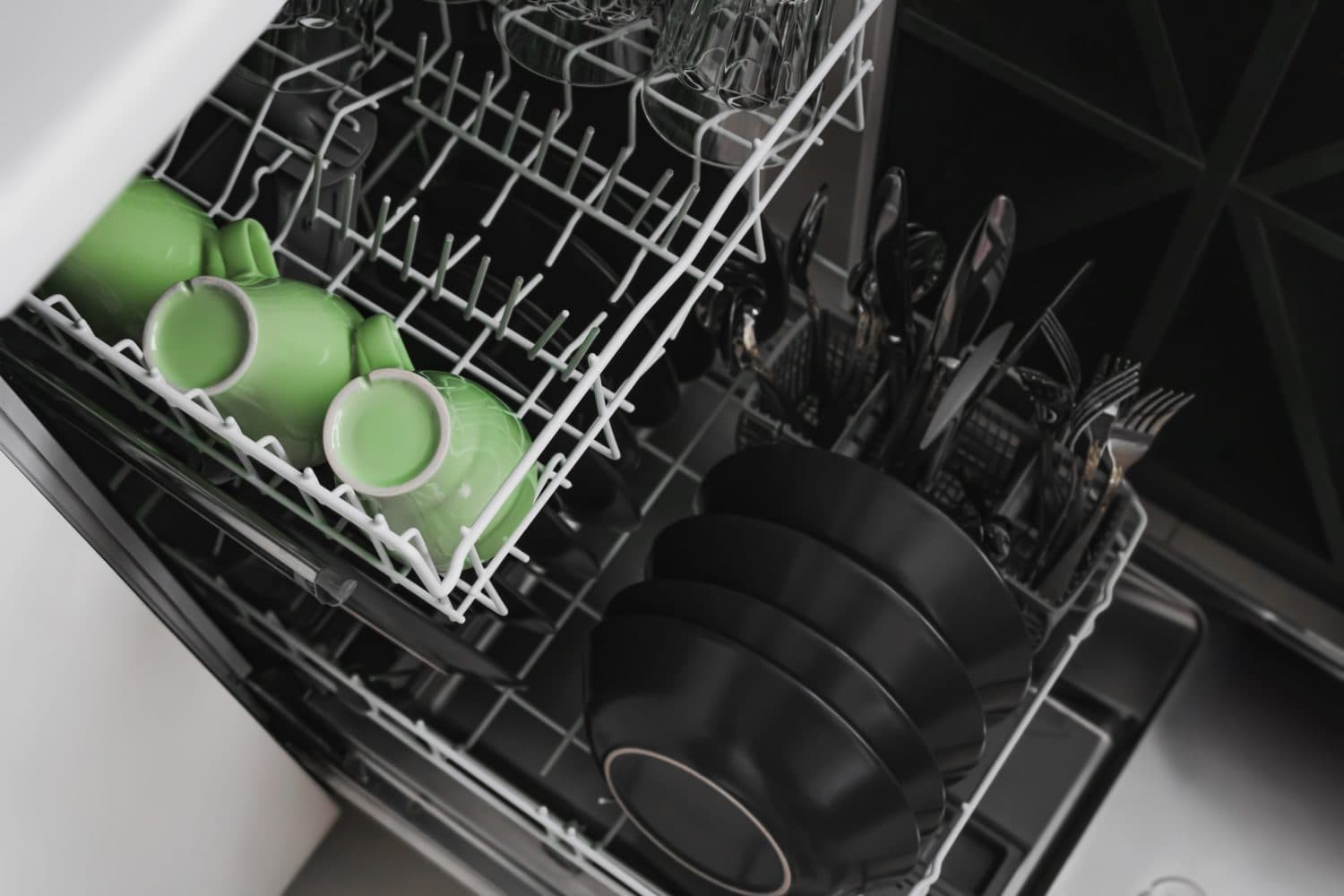 Open dishwasher with clean dishes in a bright kitchen, filled with dark plates and vibrant green cups, creating a stylish contrast. The green cups add a fresh pop to the monochrome setup.