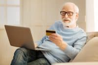Home shopping. Kind smiling male wrinkling forehead and holding card while doing online shopping