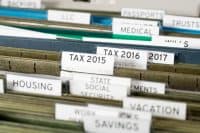 A close-up, slightly angled shot of numerous beige and blue hanging file folders in a drawer. White labels are visible on the tabs, including "BACKUPS", "LLC", "PASSPORTS", "MEDICAL", "TAX 2015", "TAX 2016", "2017", "HOUSING", "STATE SOCIAL SECURITY", "VACATION", and "SAVINGS". The focus is on the "TAX" and "HOUSING" labels.