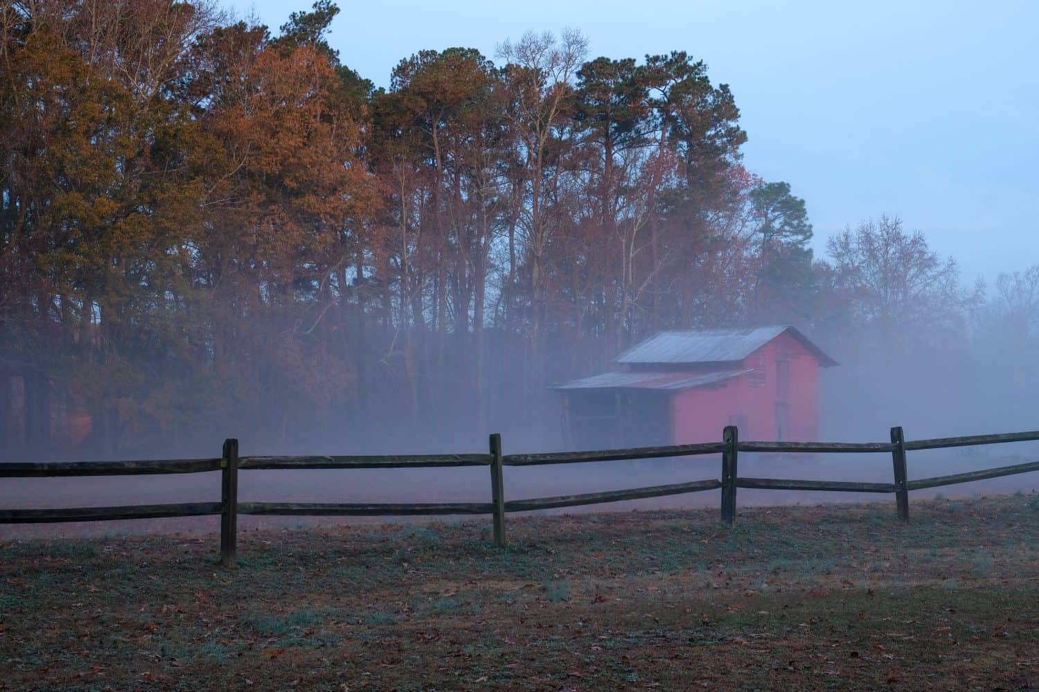 An old red barn behind a wooden fence on a foggy morning just before sunrise. Lake Benson Park in Garner North Carolina.