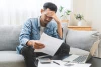 Bad financial concept, frustrated asian young man, male having debt, reading paper report from bank, stress by calculate expense from invoice or bill not able money to pay mortgage or loan, bankruptcy