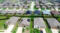 An aerial photograph captures a wide view of a modern suburban neighborhood with rows of single-family homes. Many houses feature light brown, grey, or dark blue roofs, with several clearly displaying black solar panels. The houses have well-kept green lawns and wooden fences separating properties. Cars are parked on driveways and along the streets which weave through the development. The sky is bright and clear, indicating a sunny day.