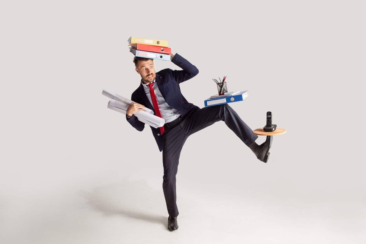 Young businessman in a suit juggling with office supplies in his office, isolated on white background. Conceptual collage with phone, folders. The business, office, work concept.