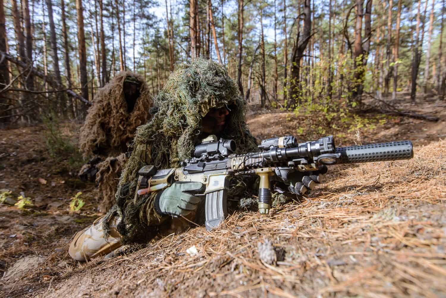Assistant sniper covers his partner during a special military operation in the forest/Snipers in camouflage suits hiding in forest