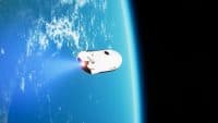 A white Intuitive Machines spacecraft with black markings and 'Intuitive' text is shown in low Earth orbit. A bright blue engine plume extends from its rear. The Earth's blue surface with white clouds fills the left side of the frame, curving towards a stark black, star-filled space on the right.
