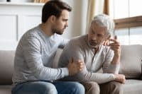 Young stressed man comforting irritated mature old father, sitting together on couch. Worried grownup son feeling sorry, asking forgiveness to angry offended unhappy middle aged hoary daddy.
