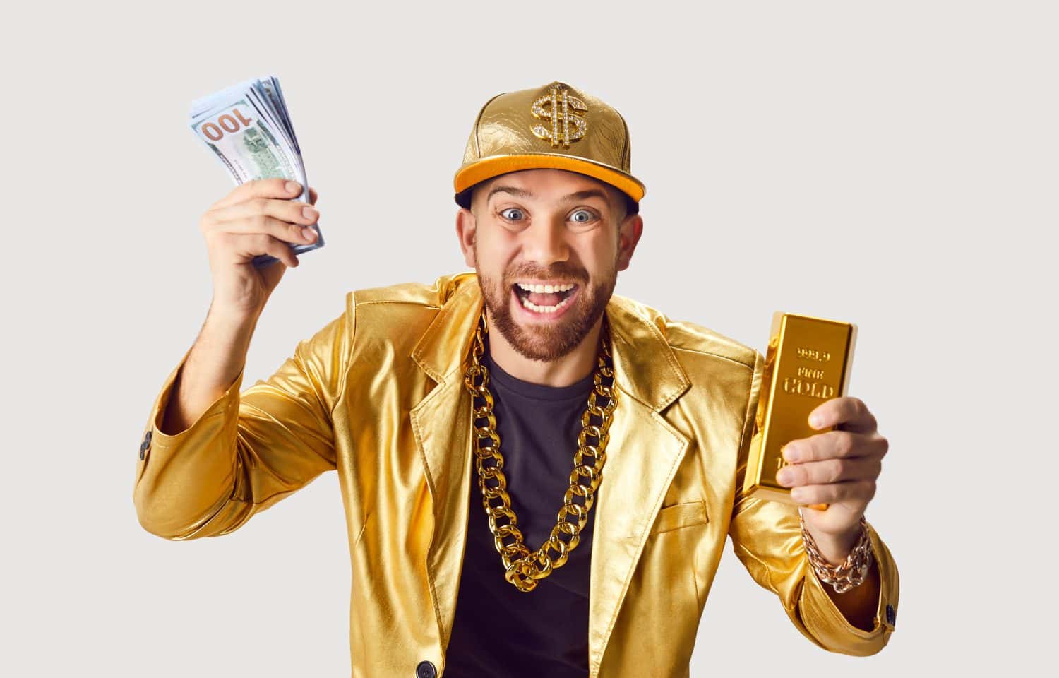 Happy excited funny young man in golden suit and cap holding money dollars cash banknotes and gold bar in hands and looking at camera isolated on white studio background. Success concept.