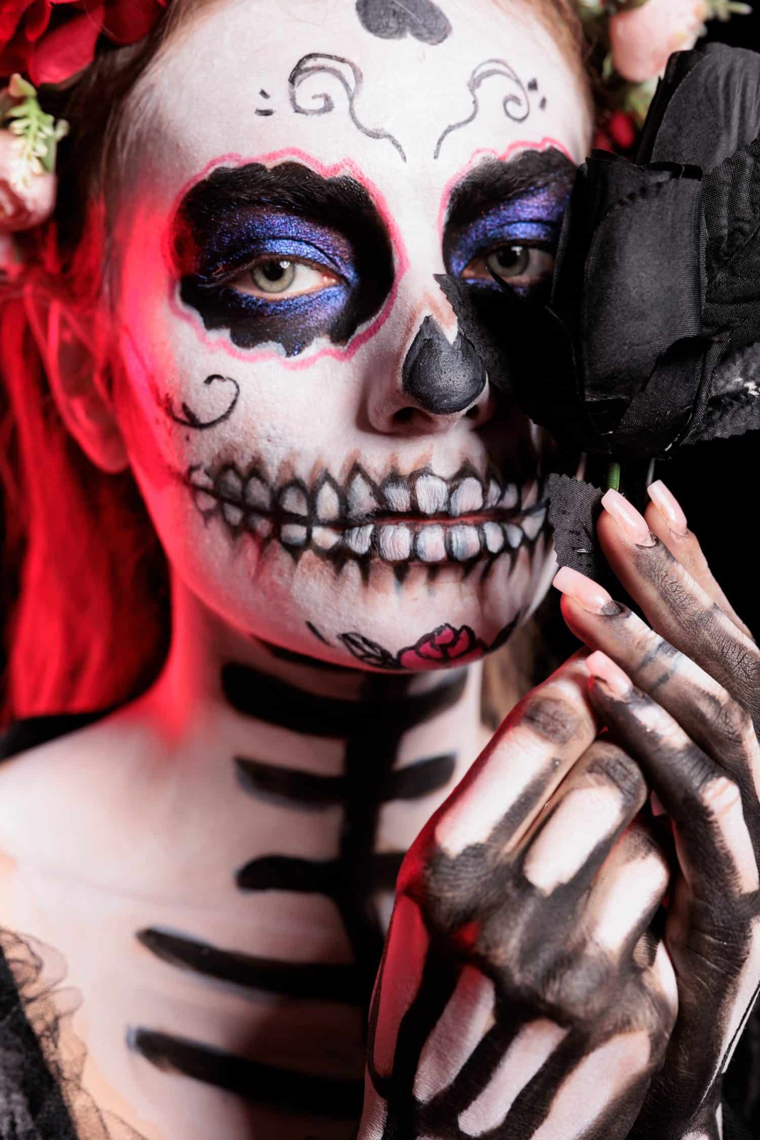 Headshot portrait of woman holding black rose participating at Dia de los Muertos party preserving cultural traditions between generations. Close up shot of lady with flower wearing la Catrina costume