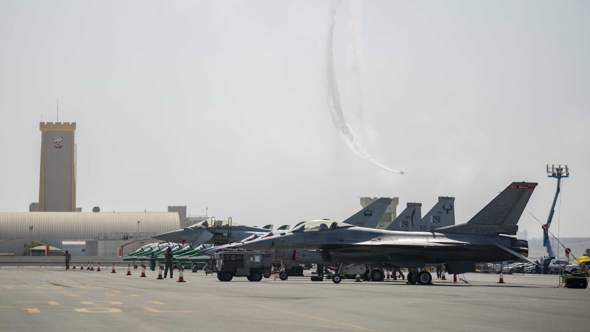 A wide shot of a flight line on a sunny day shows multiple gray fighter jets parked on the tarmac. The jets include F-16s with 'SP 480FS' on the tail and other