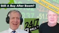 Two men, likely financial commentators, smile in front of a digital stock ticker displaying green numbers. The man on the left wears headphones, a white shirt, and a blue striped tie. The man on the right wears a black shirt. Overlays include a dark green banner on the left with white text 'Still A Buy After Boom?', a yellow diagonal banner on the top right with green text 'STOCK WATCH', and a dark green logo at the bottom center with '24/7 WALL ST'.