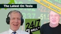 A split screen shows two smiling male financial commentators in the foreground against a blurred light green background with green and red stock market numbers. The man on the left wears a white shirt and black headphones. The man on the right wears a black shirt. Overlays include a dark green banner stating 'The Latest On Tesla,' a yellow diagonal banner reading 'STOCK WATCH,' and a green 24/7 Wall St. logo.